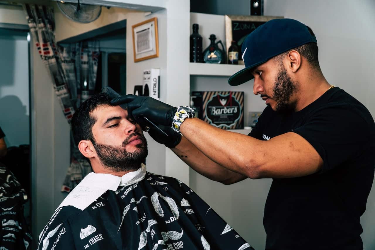 A barber wearing a black cap is shaving a man